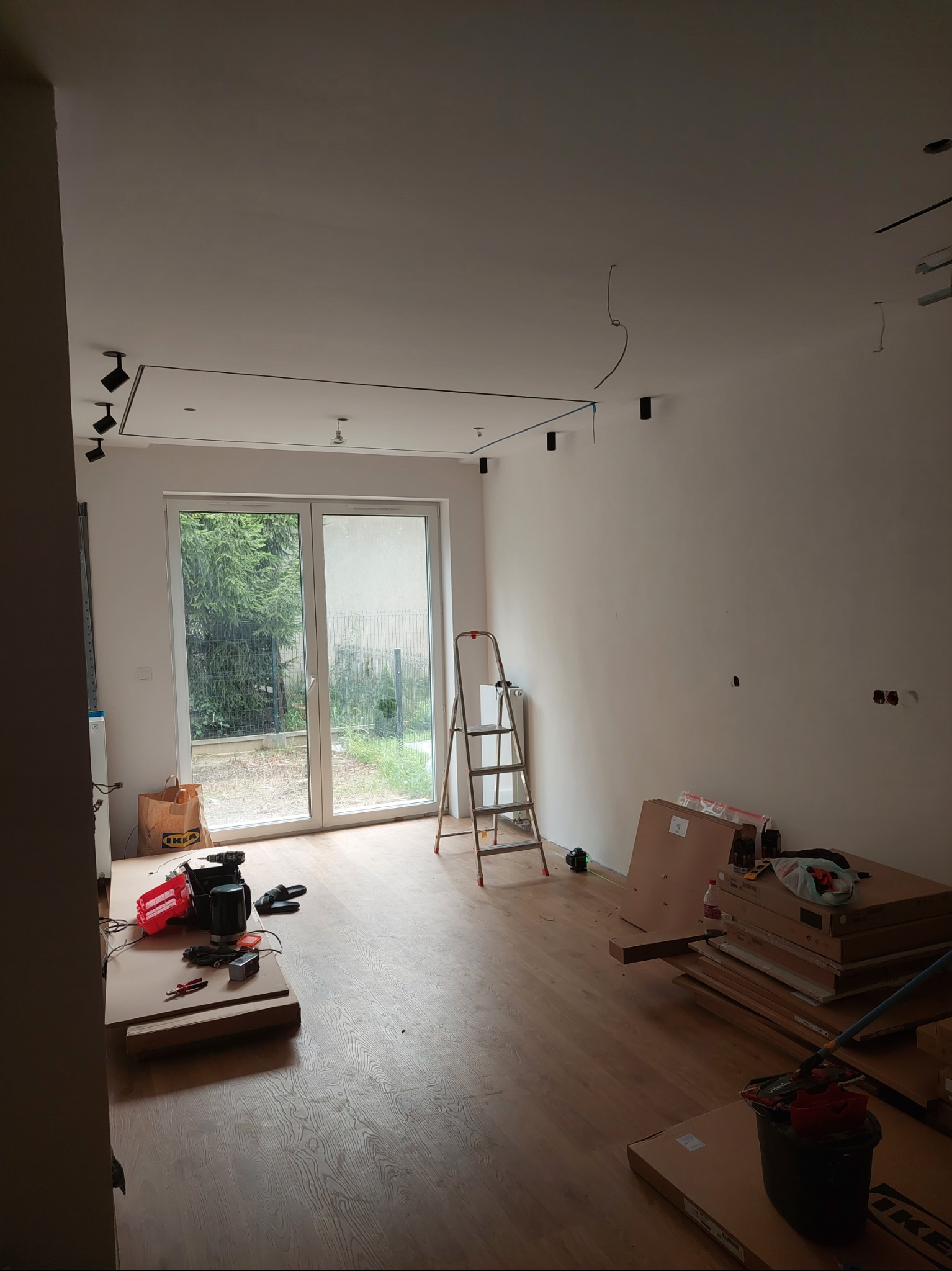 Wnętrze w trakcie prac wykończeniowych: widoczne narzędzia, drabina, podłoga i oświetlenie sufitowe w nowym mieszkaniu z wyjściem na ogród.