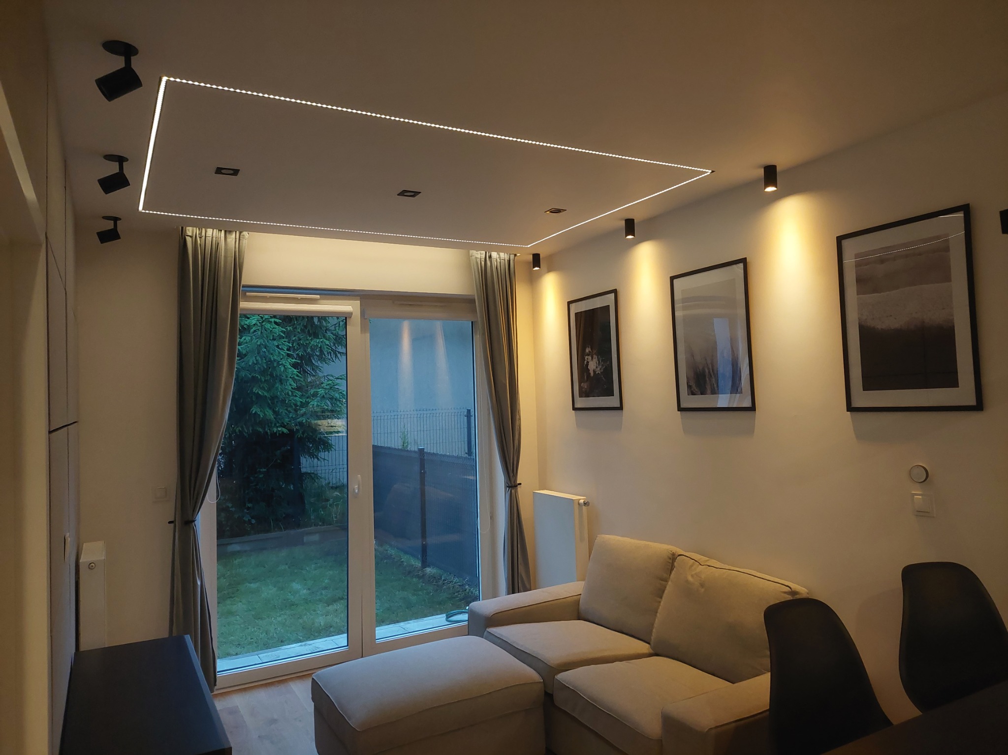 Nowoczesny salon z narożnikiem i oświetleniem LED na suficie, widok na ogród przez przeszklone drzwi balkonowe. Minimalistyczny design z czarnymi lampami punktowymi.