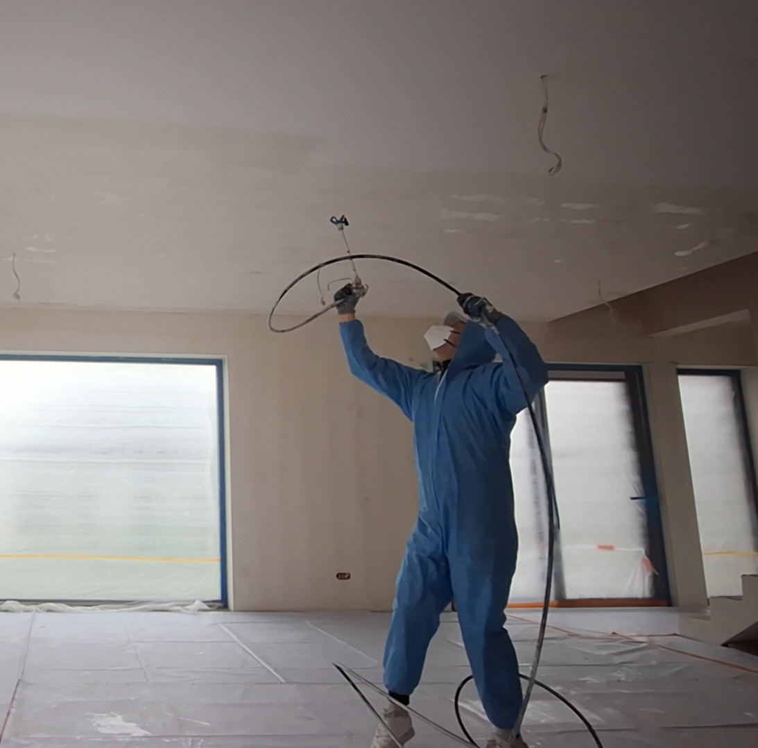 Pracownik w kombinezonie maluje sufit agregatem natryskowym w jasnym pomieszczeniu. Okna zabezpieczone folią, podłoga przykryta. Widoczne przewody i elementy oświetlenia.