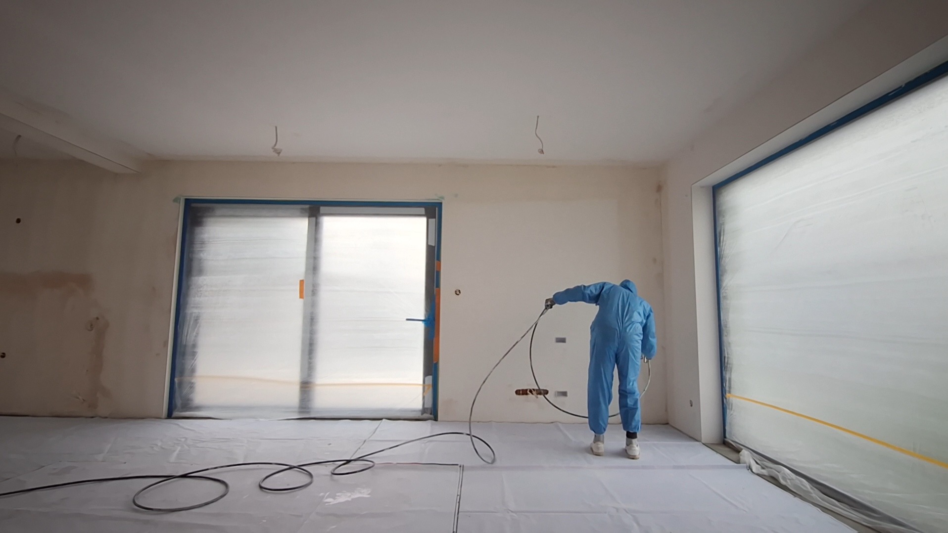 Malowanie natryskowe ścian w Ożarowie Mazowieckim. Pracownik w kombinezonie maluje ściany w pomieszczeniu zabezpieczonym folią. Widoczne okna i drzwi zabezpieczone folią.
