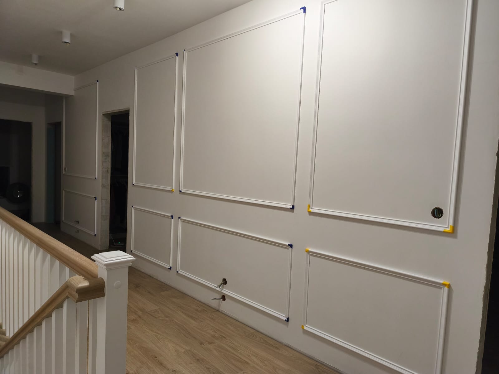 Białe ściany z dekoracyjnymi listwami w stylu klasycznym, widoczne otwory na kable i gniazdko, fragment schodów z drewnianą balustradą. Detal wykończenia wnętrza.