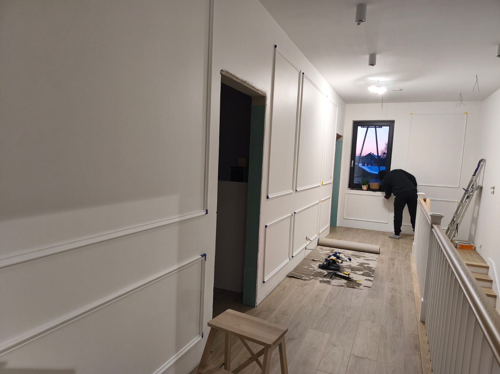 Korytarz w trakcie dekorowania sztukaterią. Widoczne listwy na ścianach, narzędzia na podłodze i osoba pracująca przy oknie. Jasne wnętrze z drewnianą podłogą.