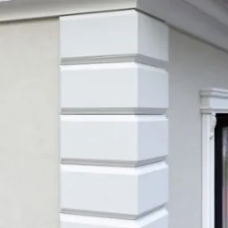 Elegancki narożnik budynku z dekoracyjnymi, białymi elementami architektonicznymi i oknami z ozdobnymi ramami. Jasna elewacja w neutralnym odcieniu.