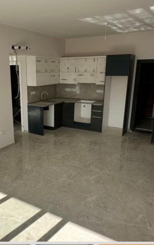 Częściowo ukończona kuchnia z szarymi szafkami i marmurową podłogą. Górne szafki jeszcze w folii ochronnej. Widok na wnętrze z połyskującą podłogą.