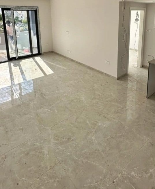 Jasna, marmurkowa podłoga z połyskiem w pustym pokoju z dużym oknem balkonowym i otwartym przejściem do kolejnego pomieszczenia. Widoczne kable.