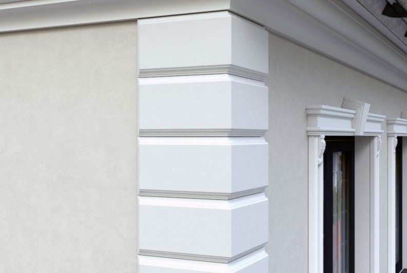 Elegancki narożnik budynku z dekoracyjnymi, białymi elementami architektonicznymi i oknami z ozdobnymi ramami. Jasna elewacja w neutralnym odcieniu.