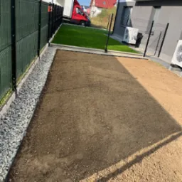 Przygotowany teren pod trawnik z rolki, widoczne obrzeża i wyrównana ziemia. W tle ogrodzenie panelowe, budynek z klimatyzacją i zaparkowany samochód dostawczy.