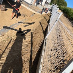 Usługi ogrodnicze Hubert Solarz - Mężczyzna z motyką przygotowuje teren pod ogrodzenie panelowe. Widoczny fragment budynku i pole kukurydzy w tle. Długi cień na ziemi.