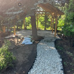 Usługi ogrodnicze Hubert Solarz - Ścieżka z białego żwiru w ogrodzie, otoczona czarnym obrzeżem, prowadząca do drewnianej altany. Worki z korą obok drzewa. Słoneczny dzień.