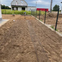 Przygotowanie podłoża pod trawnik z siatką wzmacniającą, widoczne narzędzia ogrodnicze i budynek w tle. Ujęcie z perspektywy ogrodu w trakcie zakładania.