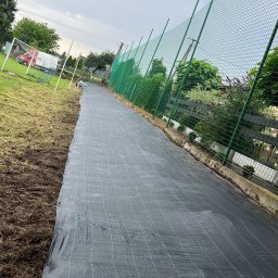 Usługi ogrodnicze Hubert Solarz - Rozłożona czarna agrowłóknina na przygotowanym terenie pod przyszły ogród, widoczne ogrodzenie z siatki i fragment boiska z bramką w tle.