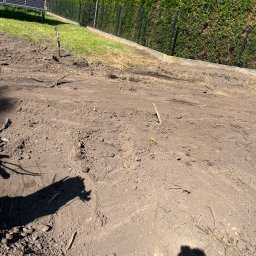 Usługi ogrodnicze Hubert Solarz - Przygotowany teren pod trawnik z widocznymi śladami po maszynach, w tle tuje i panele słoneczne. Dzień słoneczny.