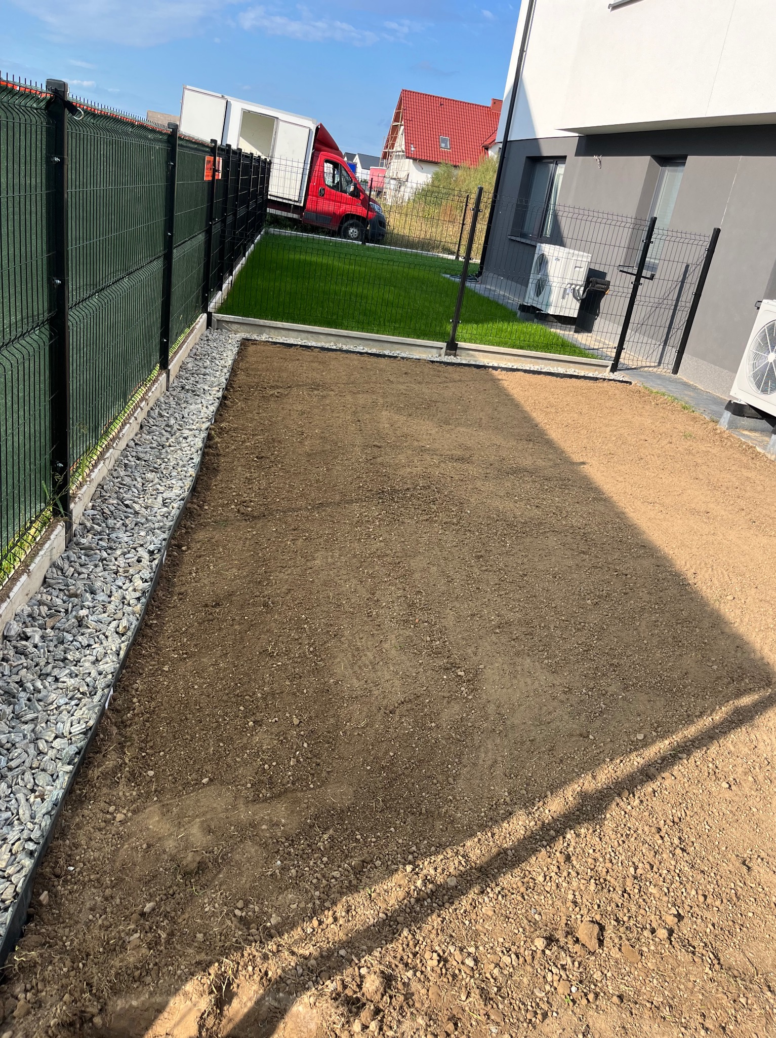 Przygotowany teren pod trawnik z rolki, widoczne obrzeża i wyrównana ziemia. W tle ogrodzenie panelowe, budynek z klimatyzacją i zaparkowany samochód dostawczy.