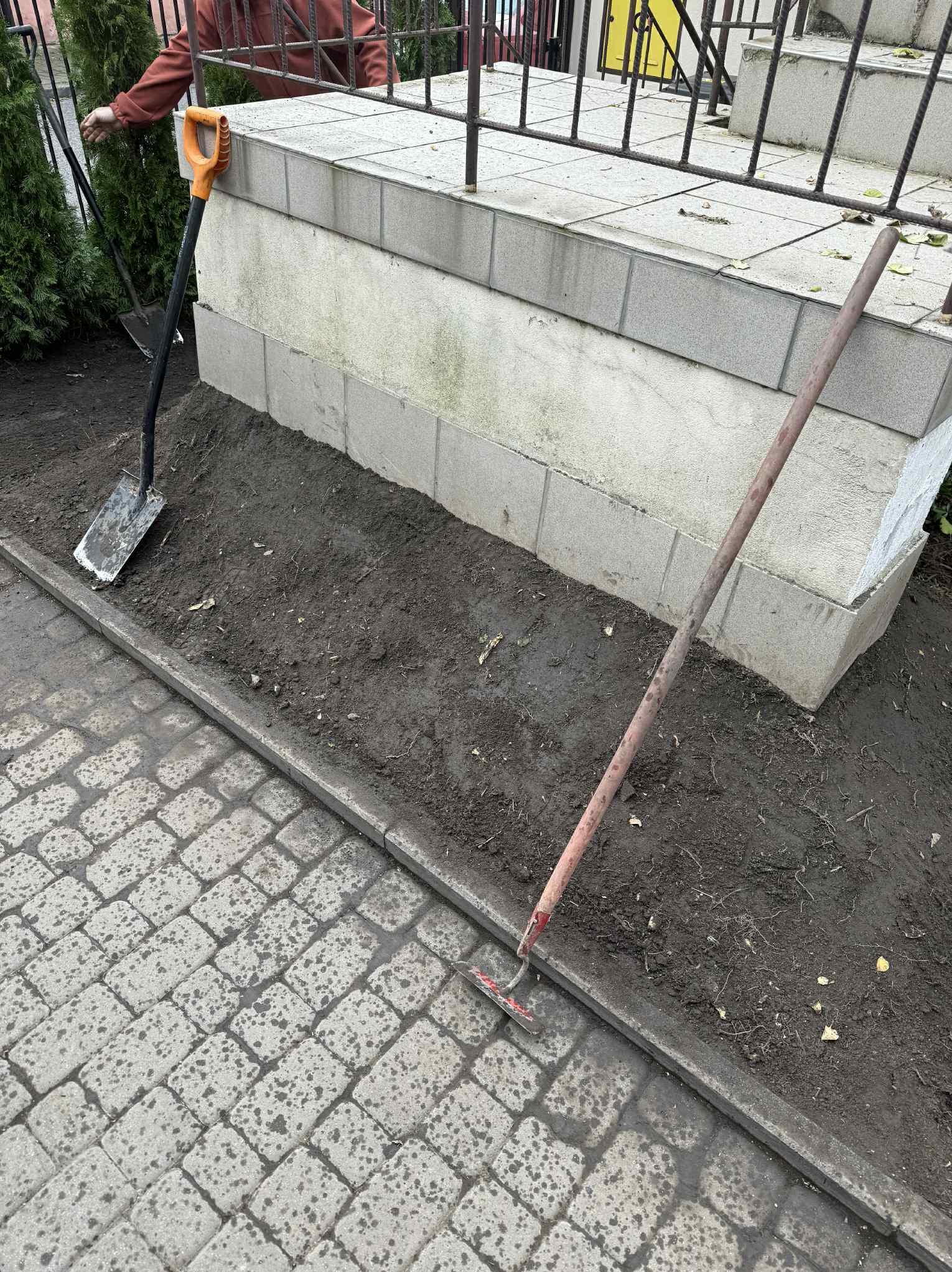 Przygotowany teren pod nasadzenia: gleba spulchniona, widoczna łopata z pomarańczowym uchwytem i grabie. Kostka brukowa obok rabaty.