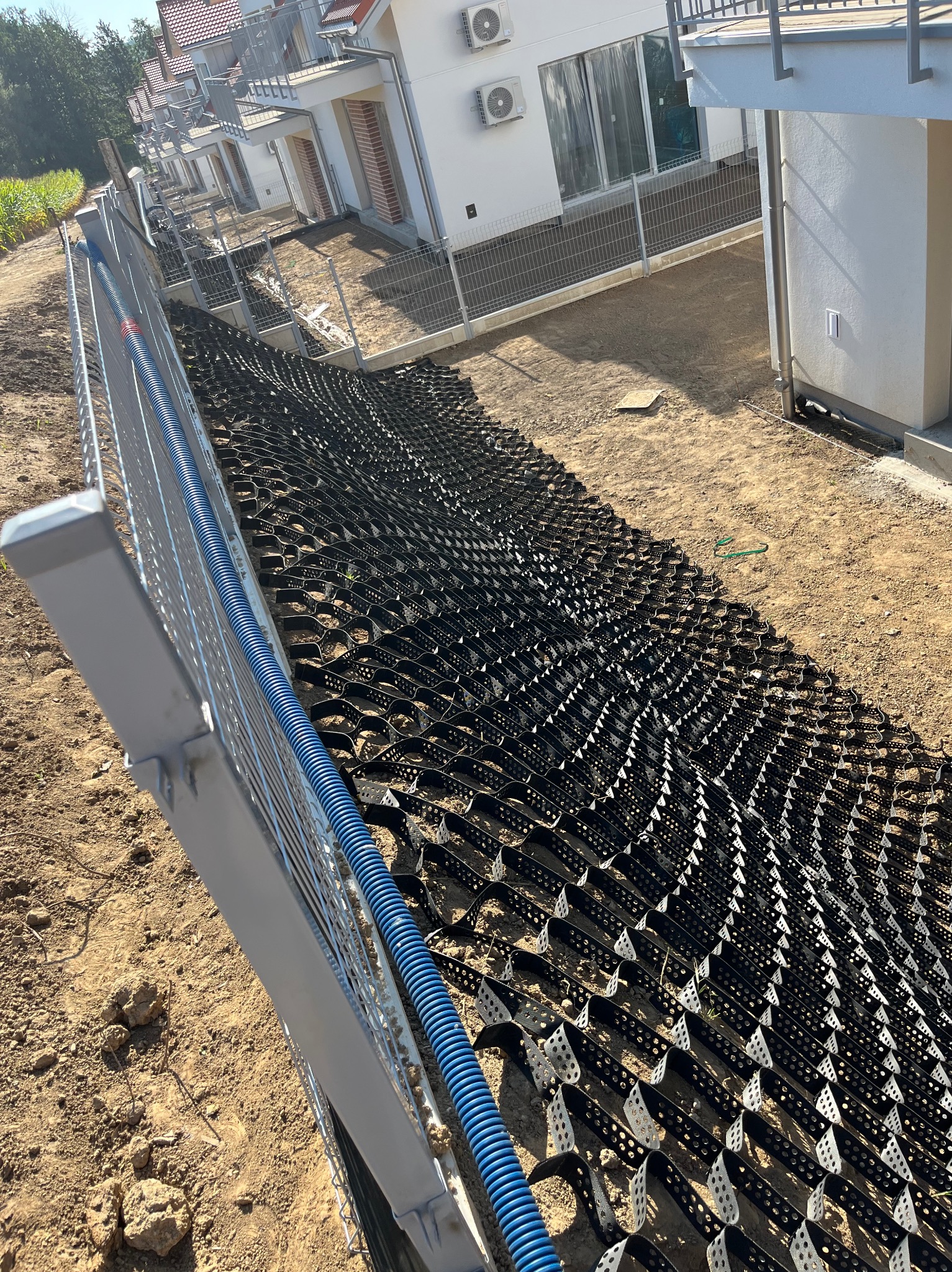 Geokratka ułożona na gruncie przy ogrodzeniu posesji. Widoczne budynki mieszkalne w tle. Grunt przygotowany pod dalsze prace.