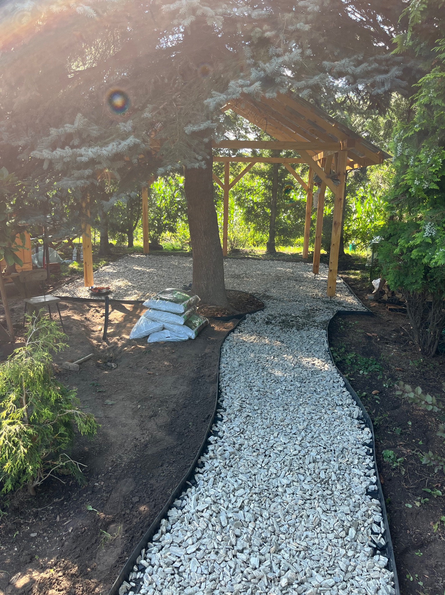 Ścieżka z białego żwiru w ogrodzie, otoczona czarnym obrzeżem, prowadząca do drewnianej altany. Worki z korą obok drzewa. Słoneczny dzień.