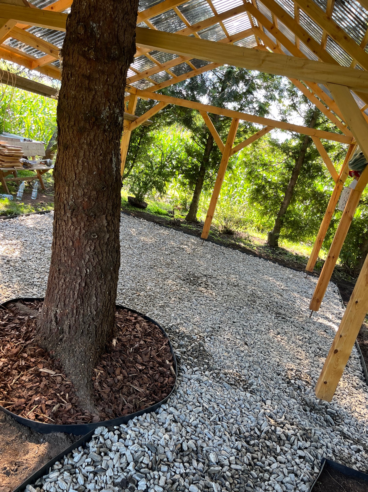 Drewniana konstrukcja altany w ogrodzie z drzewem w centrum, podsypana korą i żwirem, z widokiem na zieleń. Elementy konstrukcyjne w trakcie montażu.