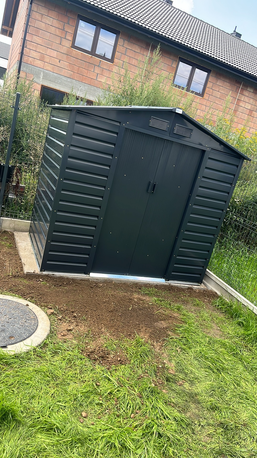 Nowoczesna, antracytowa drewutnia z metalu na betonowej wylewce, tuż obok trawnika. W tle widoczny fragment domu z cegły i okna.