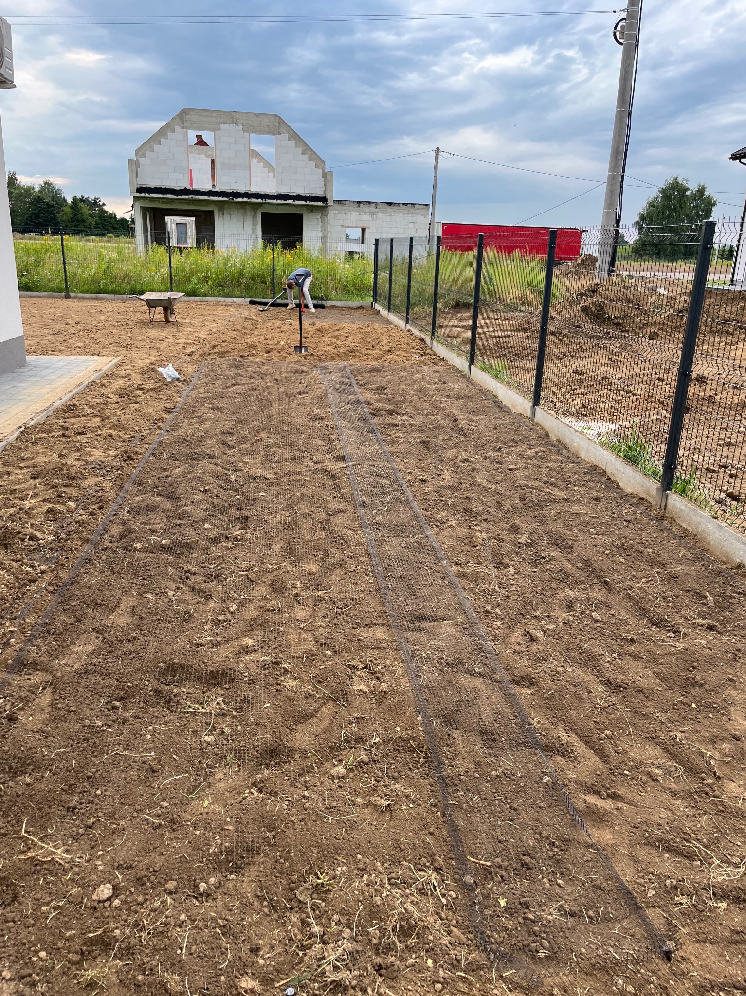 Przygotowanie podłoża pod trawnik z siatką wzmacniającą, widoczne narzędzia ogrodnicze i budynek w tle. Ujęcie z perspektywy ogrodu w trakcie zakładania.
