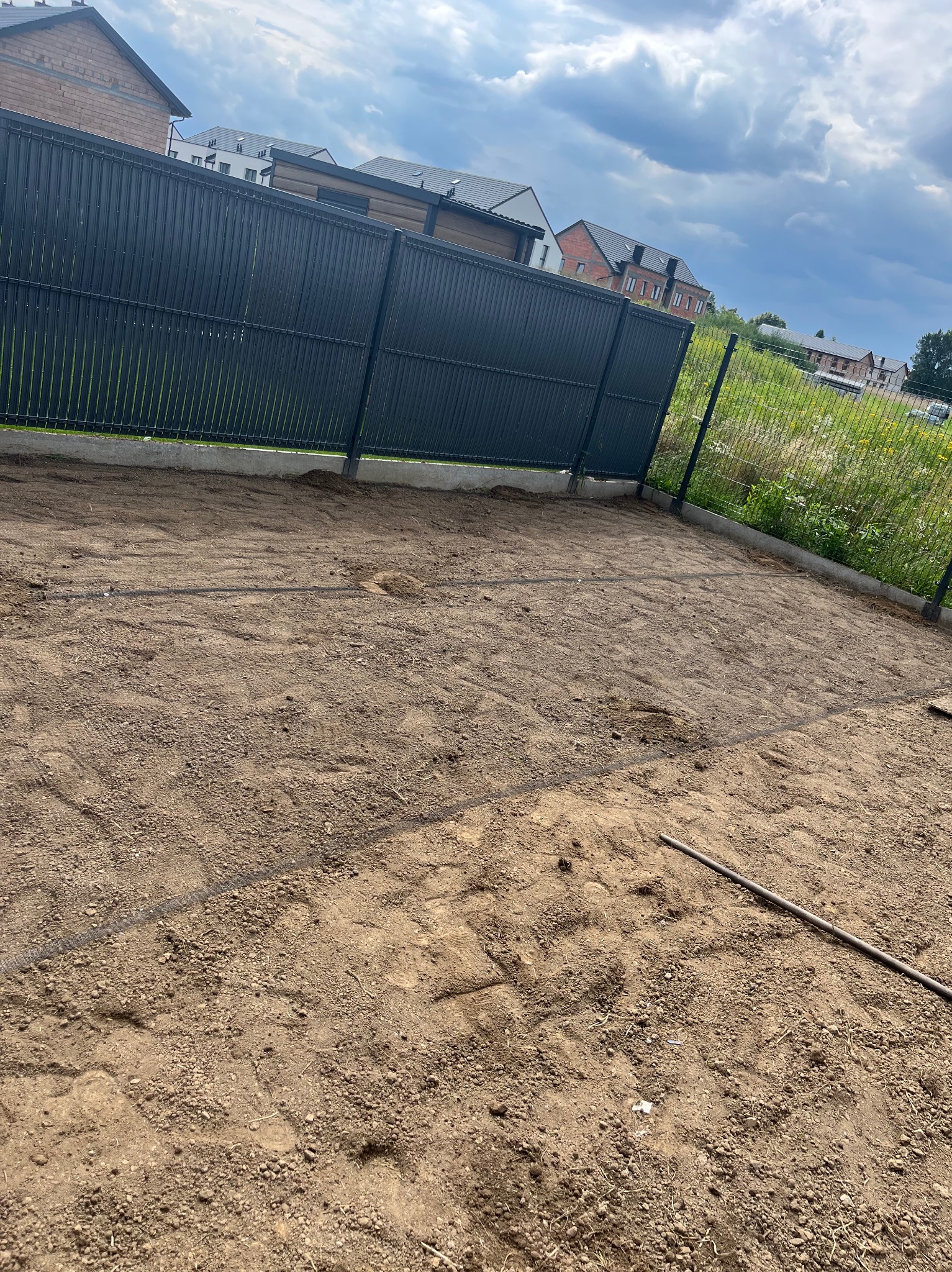 Przygotowany teren pod trawnik z rolki, widoczne ślady po maszynach i siatka. W tle metalowe ogrodzenie i zabudowa jednorodzinna. Pochmurne niebo.