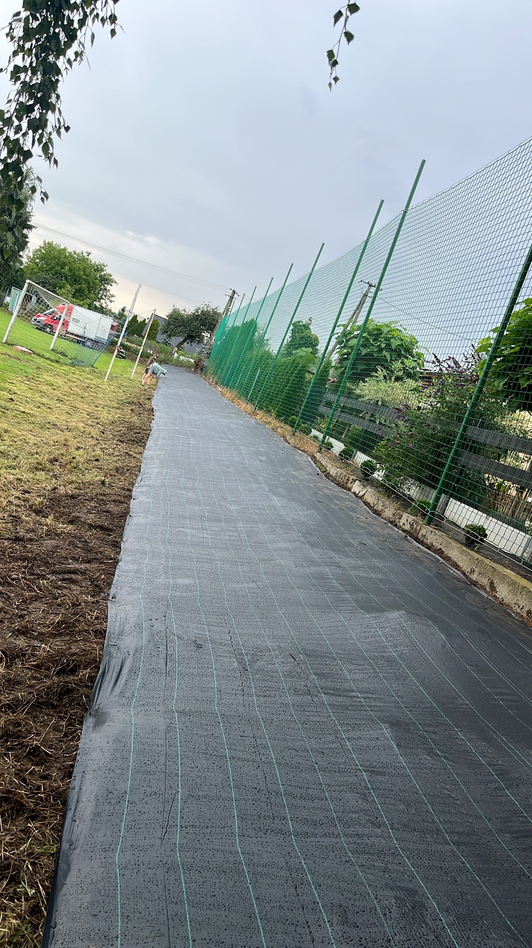 Rozłożona czarna agrowłóknina na przygotowanym terenie pod przyszły ogród, widoczne ogrodzenie z siatki i fragment boiska z bramką w tle.