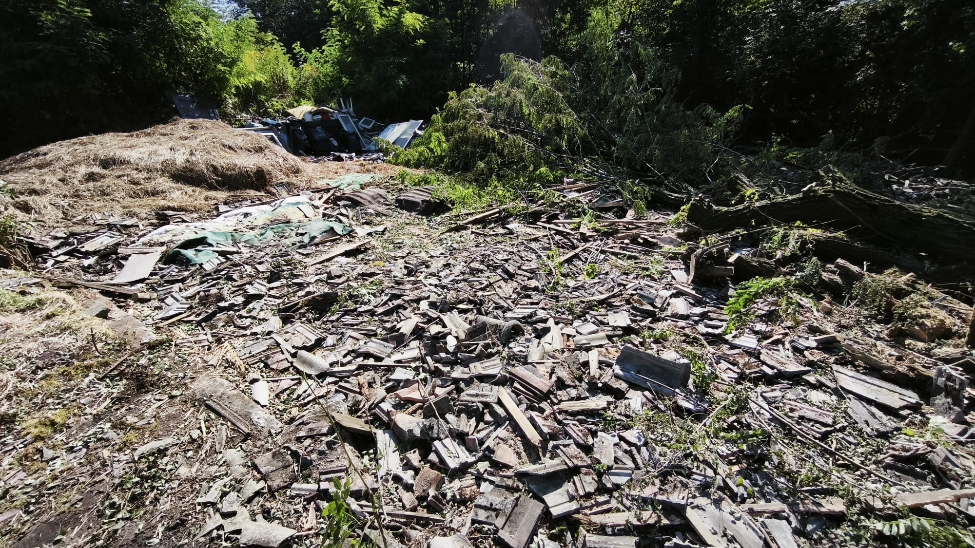 Sterta gruzu, potłuczonych dachówek i odpadów budowlanych na zewnątrz w otoczeniu drzew. Widoczne fragmenty okien. Prawdopodobnie po wyburzeniu lub remoncie.