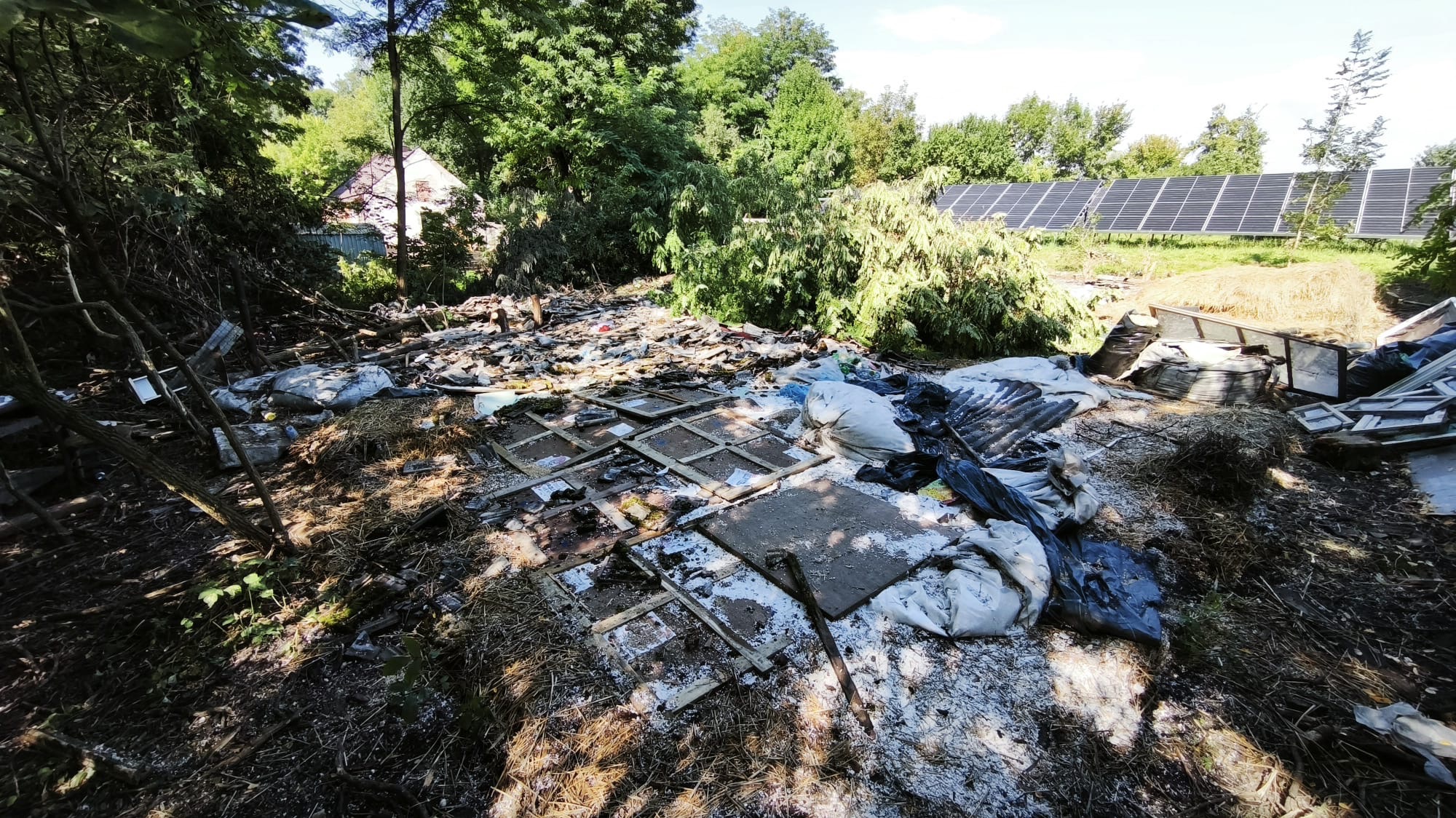 Teren po wycince drzew z pozostałościami gałęzi, pni i odpadów budowlanych. W tle widoczne panele słoneczne i zieleń.