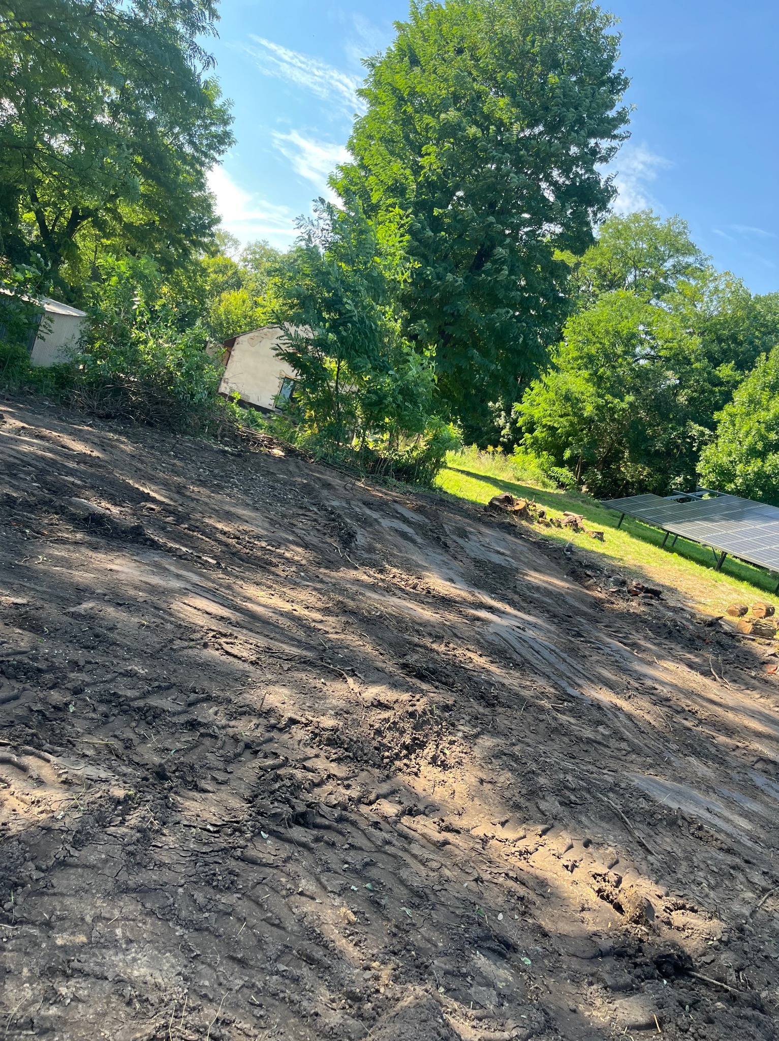 Ukształtowany teren pod budowę z widocznymi śladami opon, w tle zieleń drzew i zabudowania. Słoneczny dzień, błękitne niebo z delikatnymi obłokami.