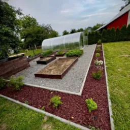 Ogród z rabatami obrzeżonymi kostką, szarą ścieżką i tunelem foliowym w tle. Dekoracyjna kora rozsypana na rabatach.