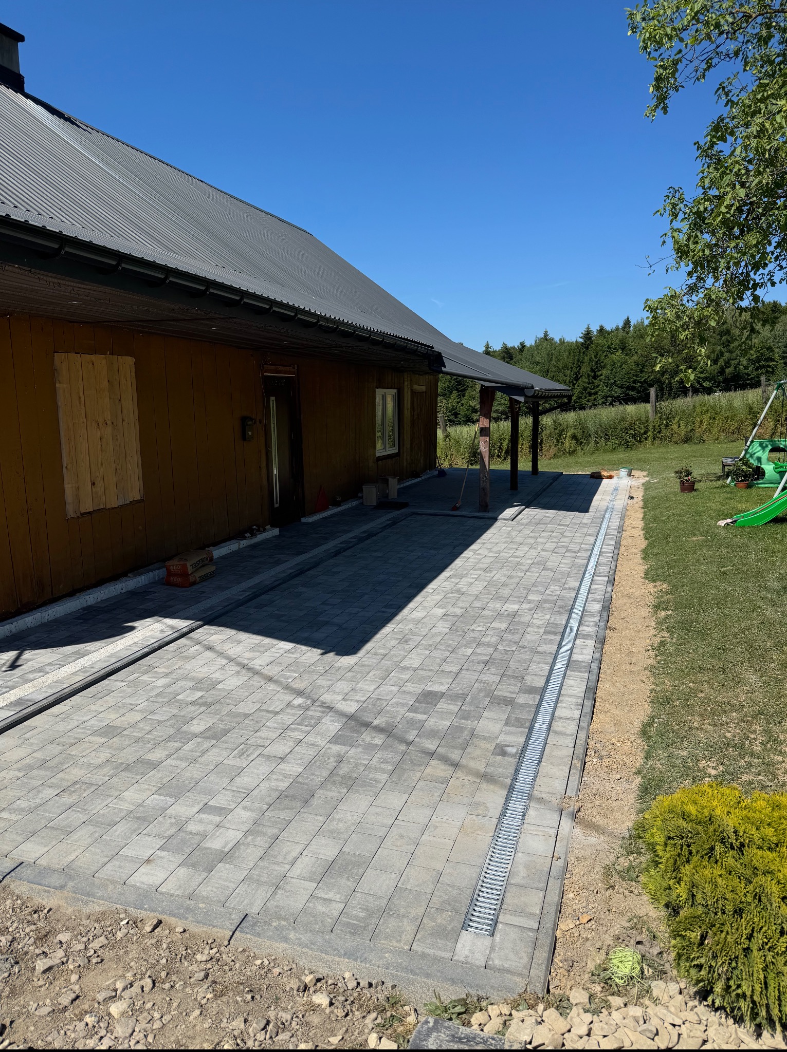 Ułożona szara kostka brukowa na tarasie domu z metalowym odwodnieniem liniowym. Widoczne worki z materiałem budowlanym i fragment ogrodu.