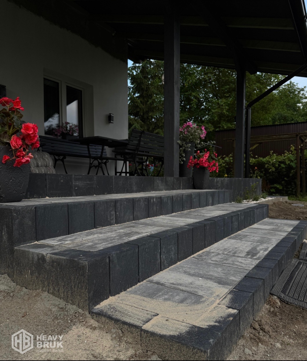 Schody z kostki brukowej w odcieniach szarości prowadzą do domu z tarasem. Kwiaty w donicach dodają koloru. Widoczny fragment ogrodu.