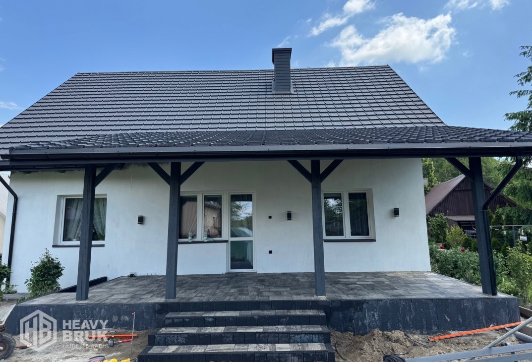 Nowoczesny taras z ciemnej kostki brukowej przed domem z białymi ścianami i ciemnym dachem. Widoczne schody z kostki i elementy wykończeniowe.