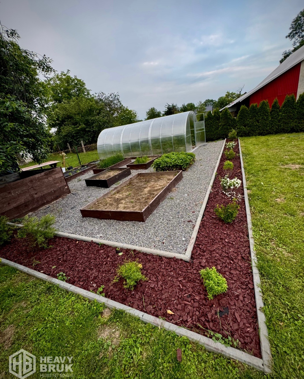Ogród z rabatami obrzeżonymi kostką, szarą ścieżką i tunelem foliowym w tle. Dekoracyjna kora rozsypana na rabatach.
