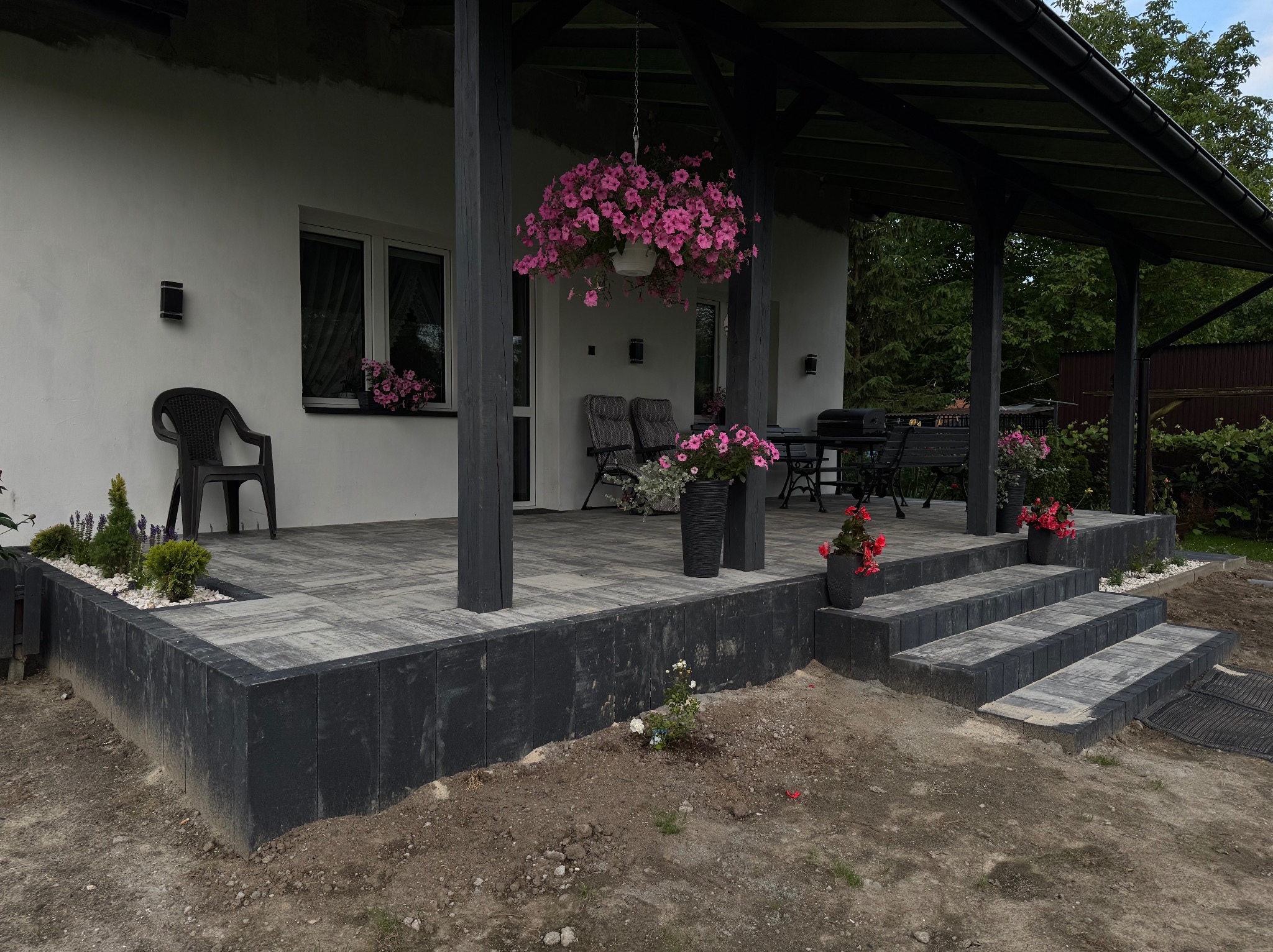 Zadaszony taras z kostki brukowej w odcieniach szarości, z czarnymi obrzeżami i schodami. Donice z kwiatami i wiszące petunie dodają koloru.