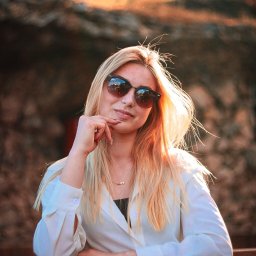 Filip Błaszczyk-Szafraniec - Portret kobiety w okularach przeciwsłonecznych, opierającej się o drewnianą barierkę, z rozproszonym światłem słonecznym w tle. Ujęcie z dołu.