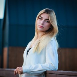 Filip Błaszczyk-Szafraniec - Portret młodej kobiety z blond włosami i niebieskimi oczami, opierającej się o drewnianą barierkę na tle ciemnoniebieskiego budynku.