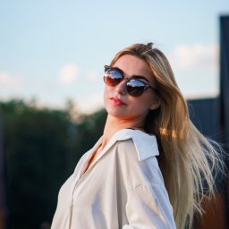 Filip Błaszczyk-Szafraniec - Portret młodej kobiety w okularach przeciwsłonecznych, z rozwianymi blond włosami, opierającej się o barierkę na tle nieba i zieleni. Ubrana w białą koszulę.