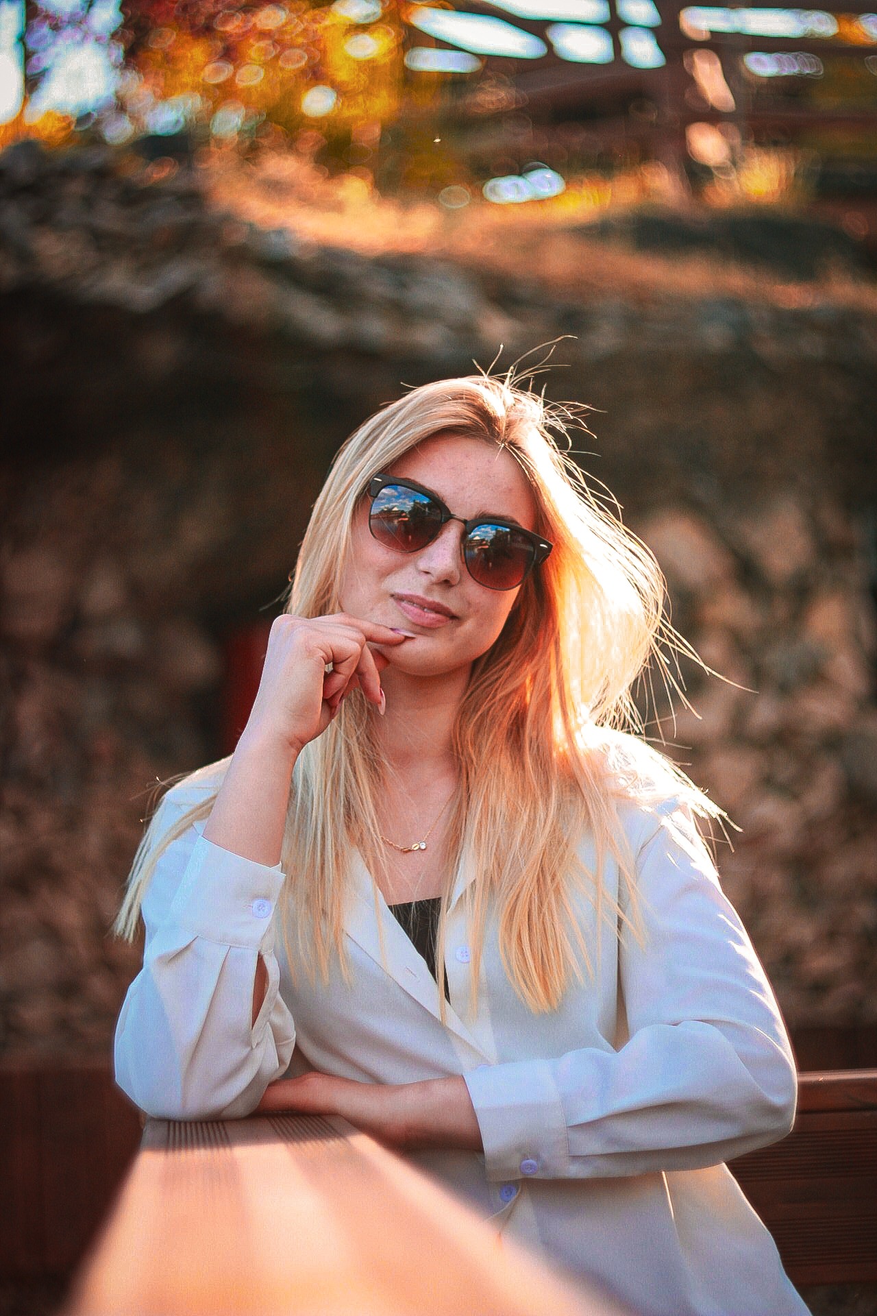 Portret kobiety w okularach przeciwsłonecznych, opierającej się o drewnianą barierkę, z rozproszonym światłem słonecznym w tle. Ujęcie z dołu.