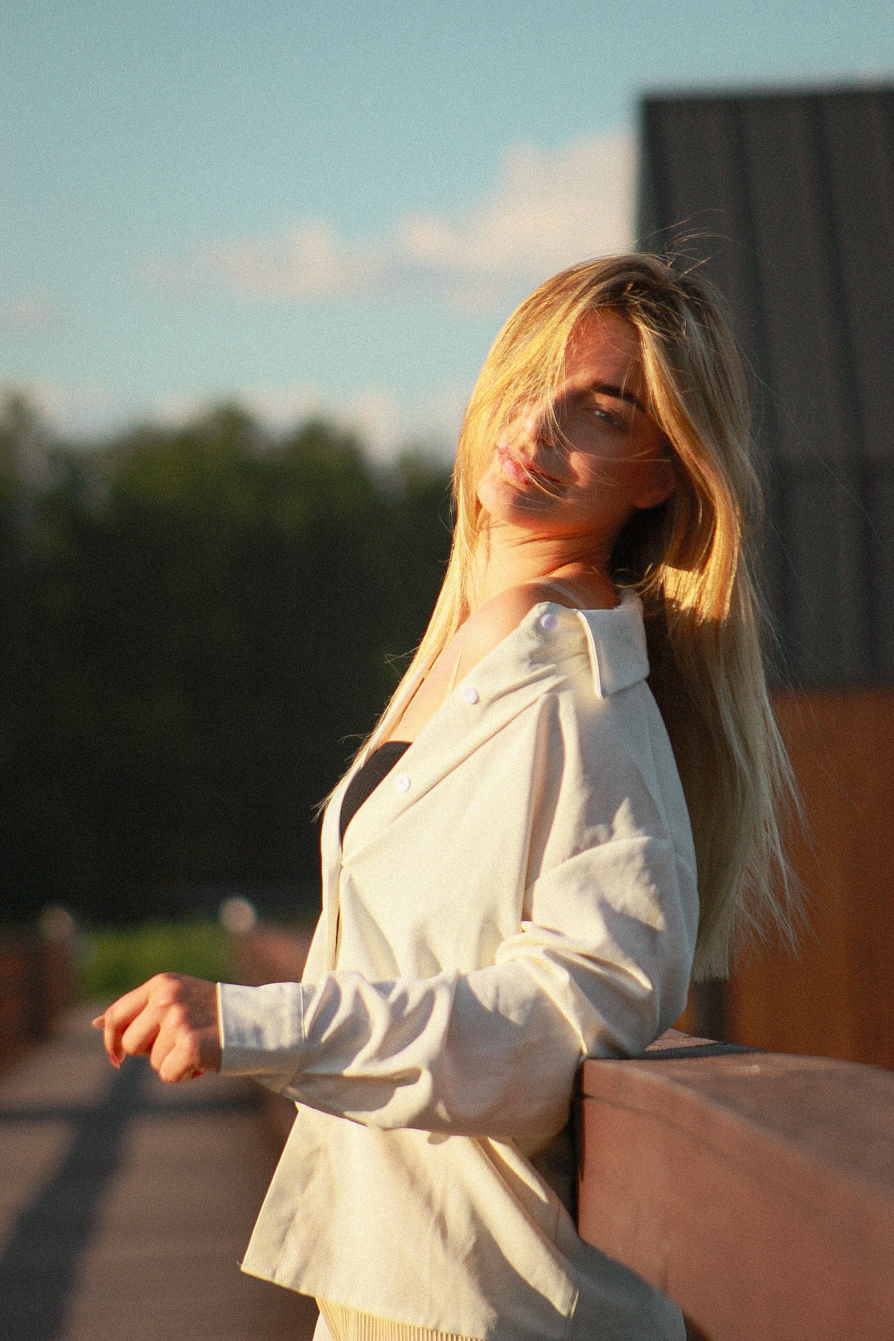 Portret blondwłosej kobiety w białej koszuli, opierającej się o brązową barierkę, z zielonym tłem i błękitnym niebem w tle, oświetlonej ciepłym światłem zachodzącego słońca.