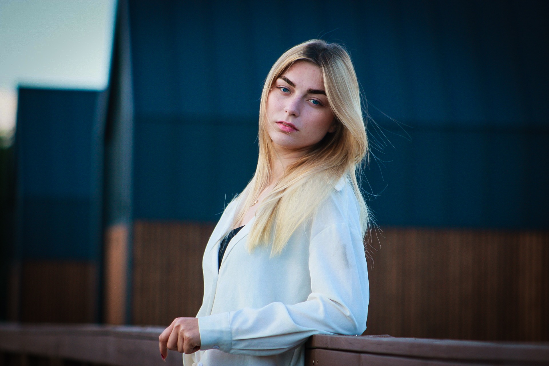 Portret młodej kobiety z blond włosami i niebieskimi oczami, opierającej się o drewnianą barierkę na tle ciemnoniebieskiego budynku.