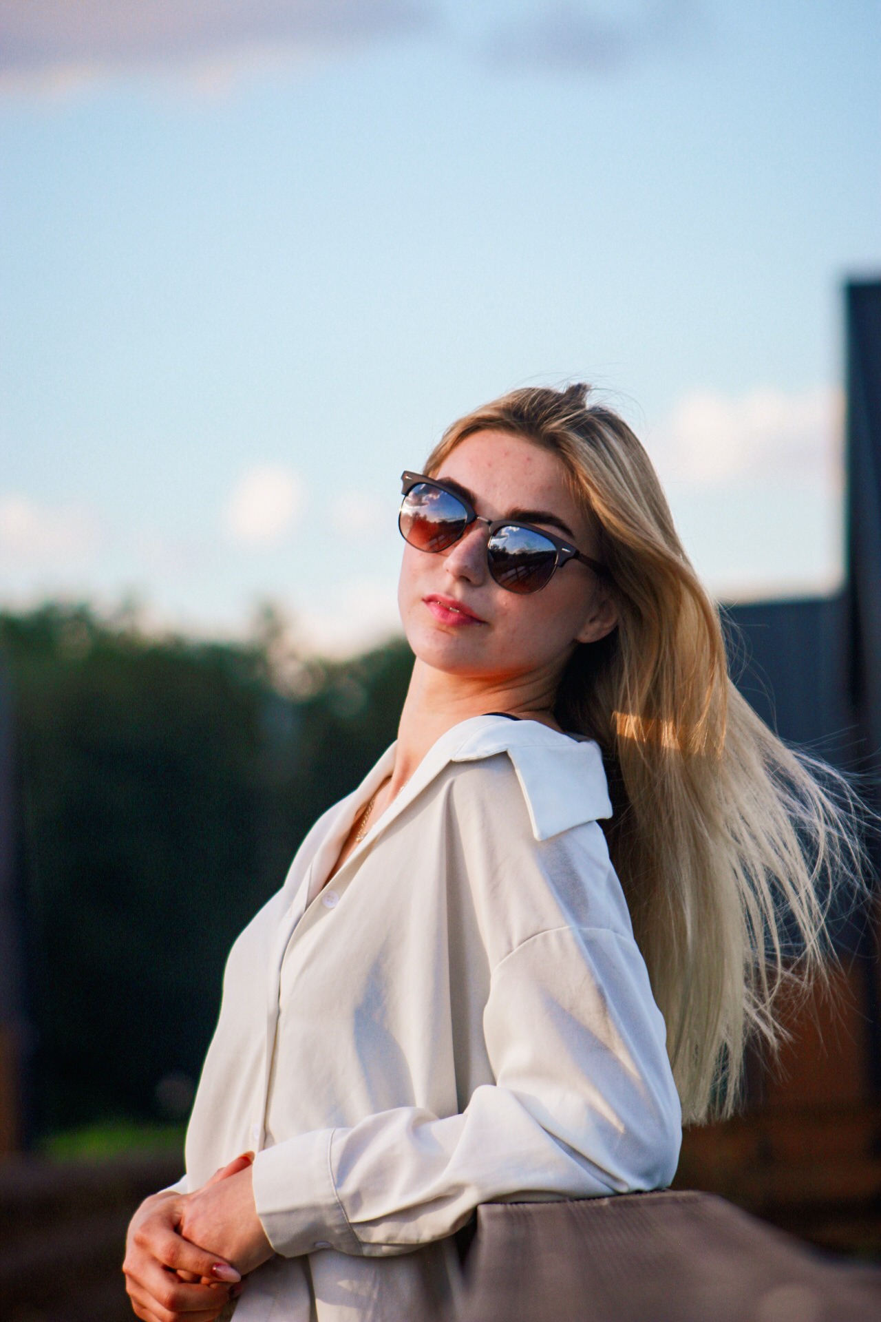 Portret młodej kobiety w okularach przeciwsłonecznych, z rozwianymi blond włosami, opierającej się o barierkę na tle nieba i zieleni. Ubrana w białą koszulę.