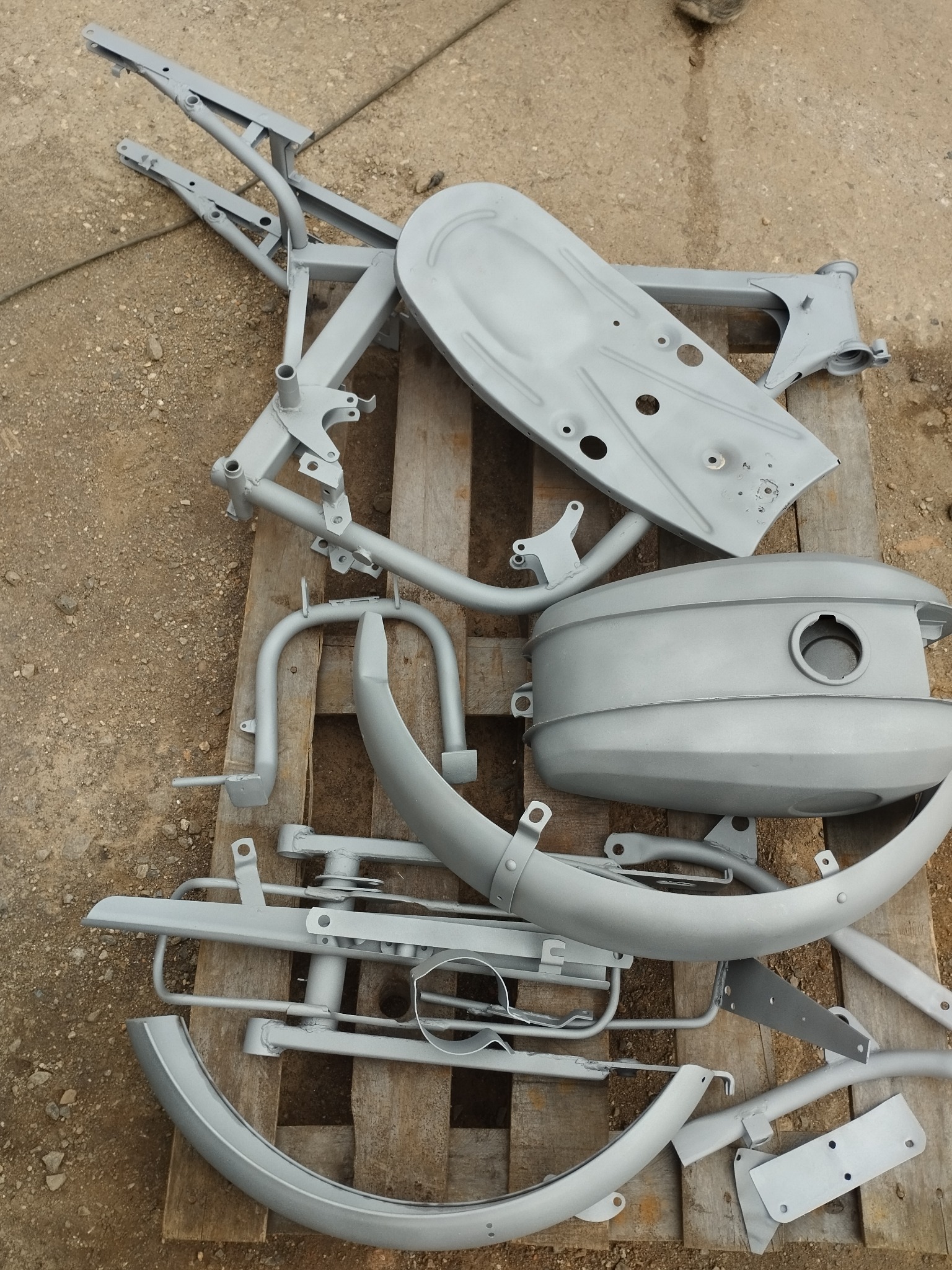 Elementy motocykla po obróbce, leżące na palecie. Widoczna rama, błotniki i zbiornik paliwa, pokryte świeżą, szarą farbą. Przygotowane do dalszego montażu.