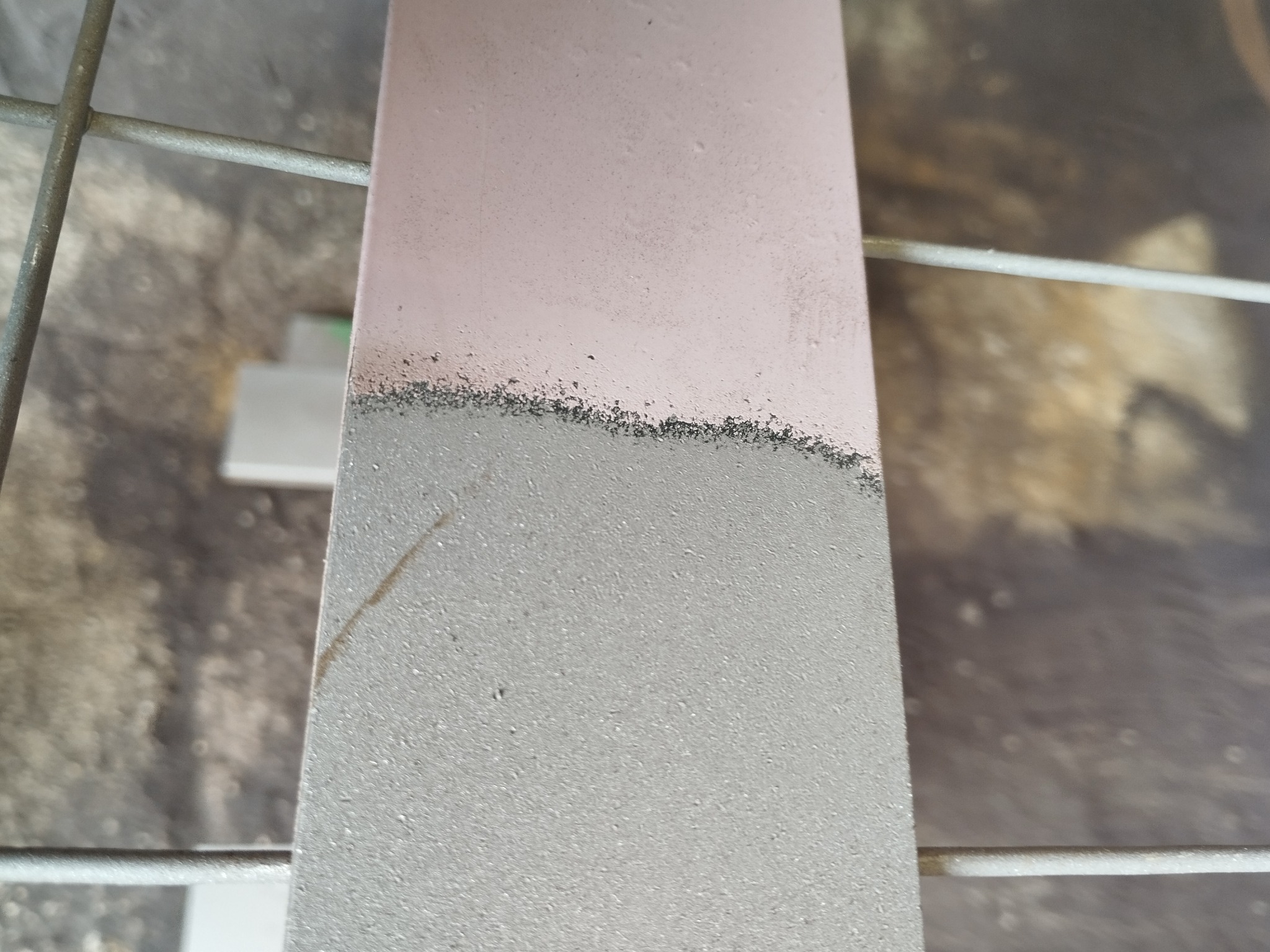 Metalowy element, częściowo oczyszczony z farby, z widoczną różnicą w strukturze powierzchni. Widoczna siatka i fragmenty podłoża w tle.