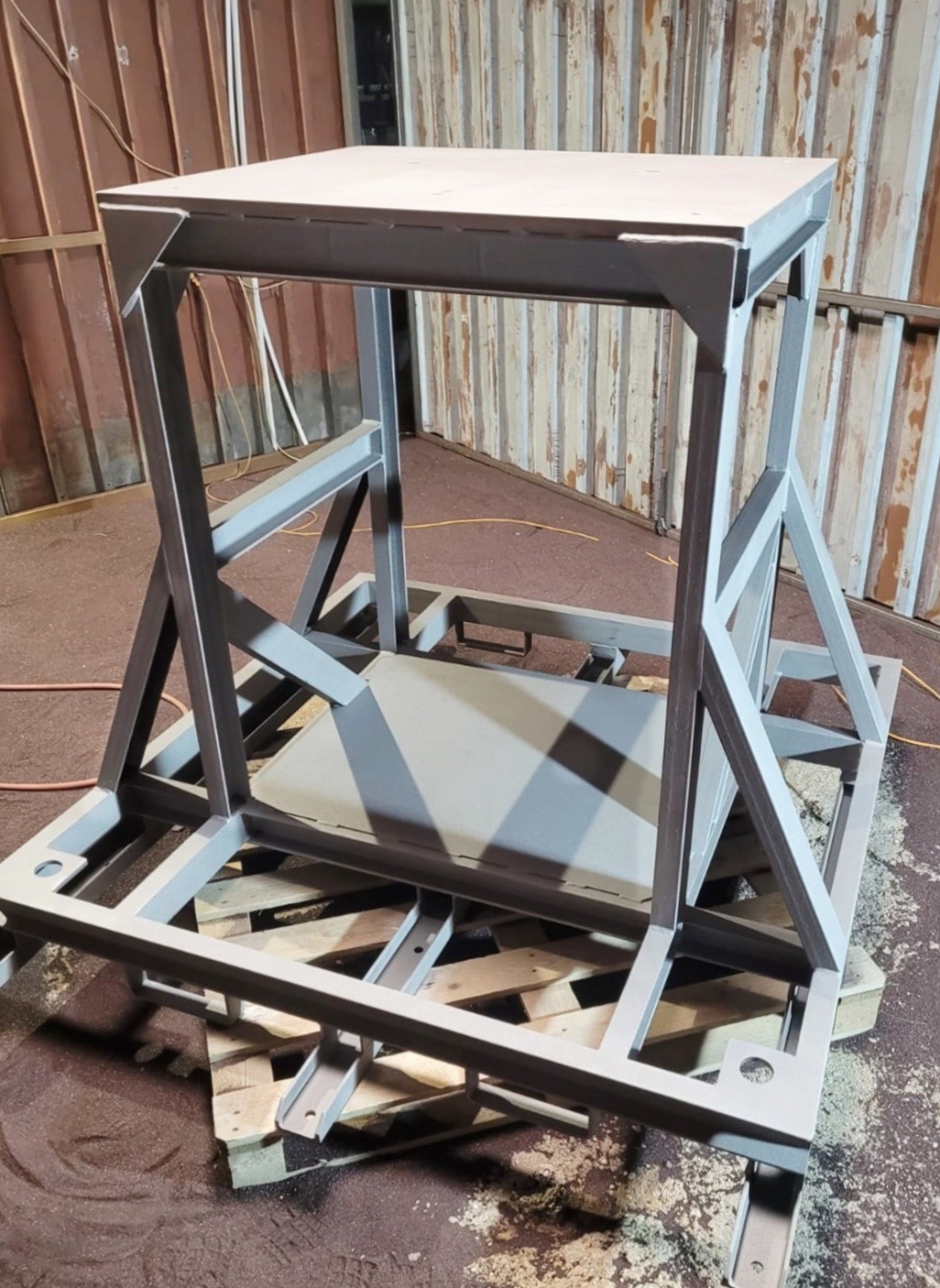 Szara, metalowa konstrukcja po obróbce, umieszczona na palecie. Widoczne spawy i elementy konstrukcyjne. Tło stanowią blaszane ściany.