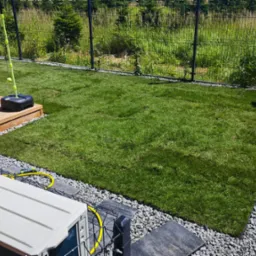 Świeżo ułożona, prostokątna trawa z rolki otoczona szarym żwirem, z widocznym fragmentem tarasu i czarnym ogrodzeniem panelowym w tle, w słoneczny dzień.