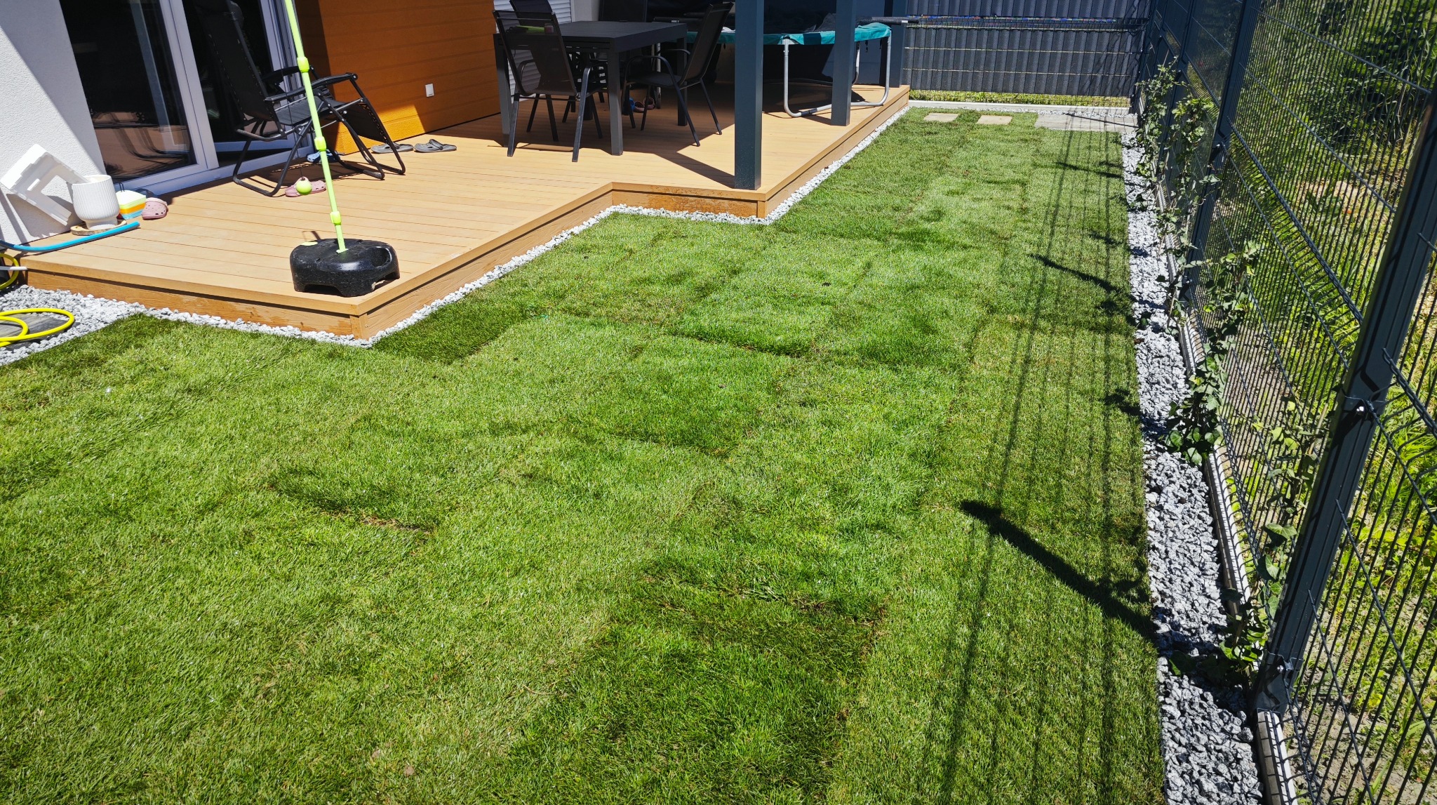 Świeżo ułożona trawa z rolki przylega do drewnianego tarasu z białymi kamykami, obok metalowe ogrodzenie z kamiennym obramowaniem. Widok z góry na równy trawnik.