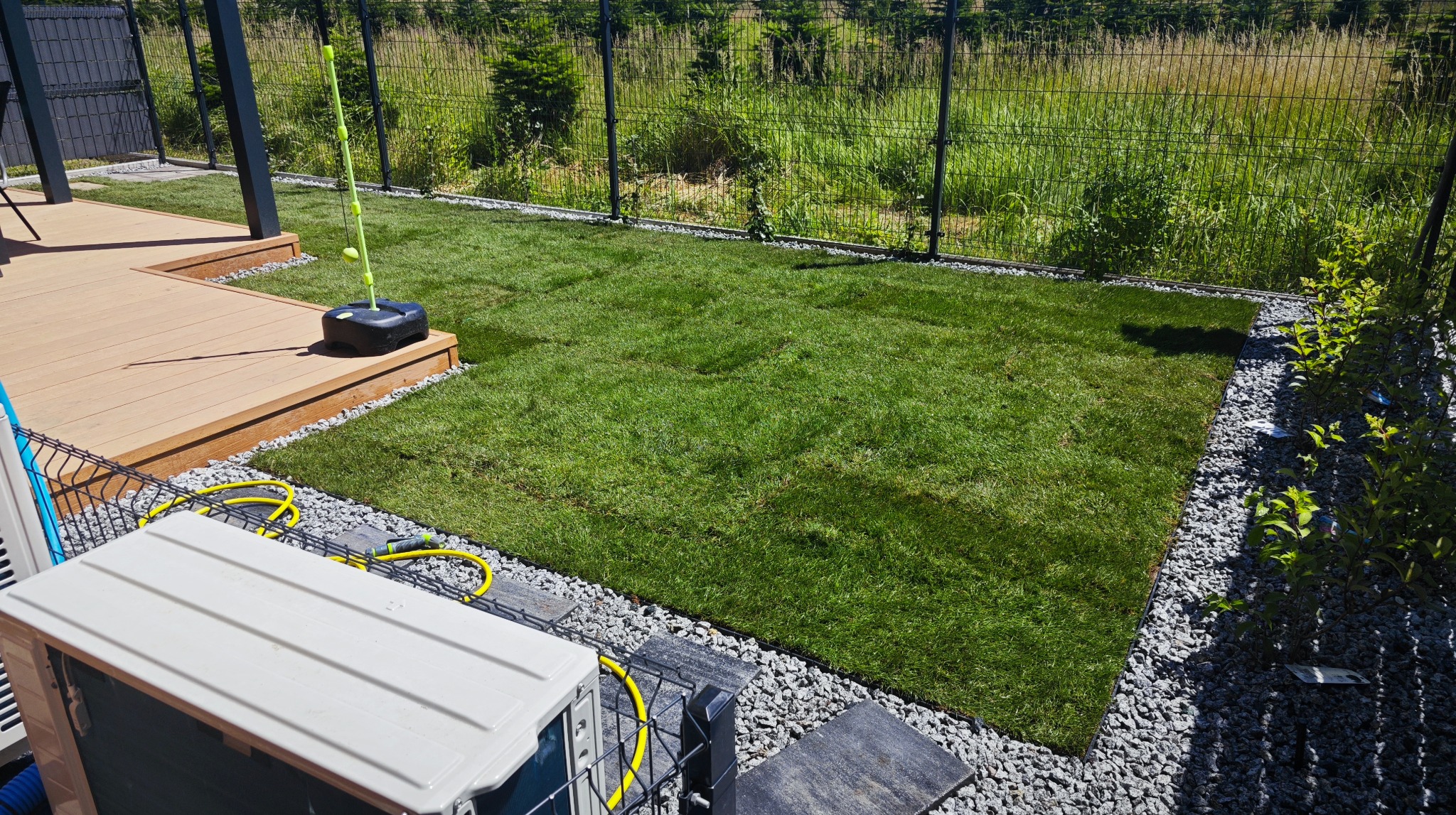 Świeżo ułożona, prostokątna trawa z rolki otoczona szarym żwirem, z widocznym fragmentem tarasu i czarnym ogrodzeniem panelowym w tle, w słoneczny dzień.