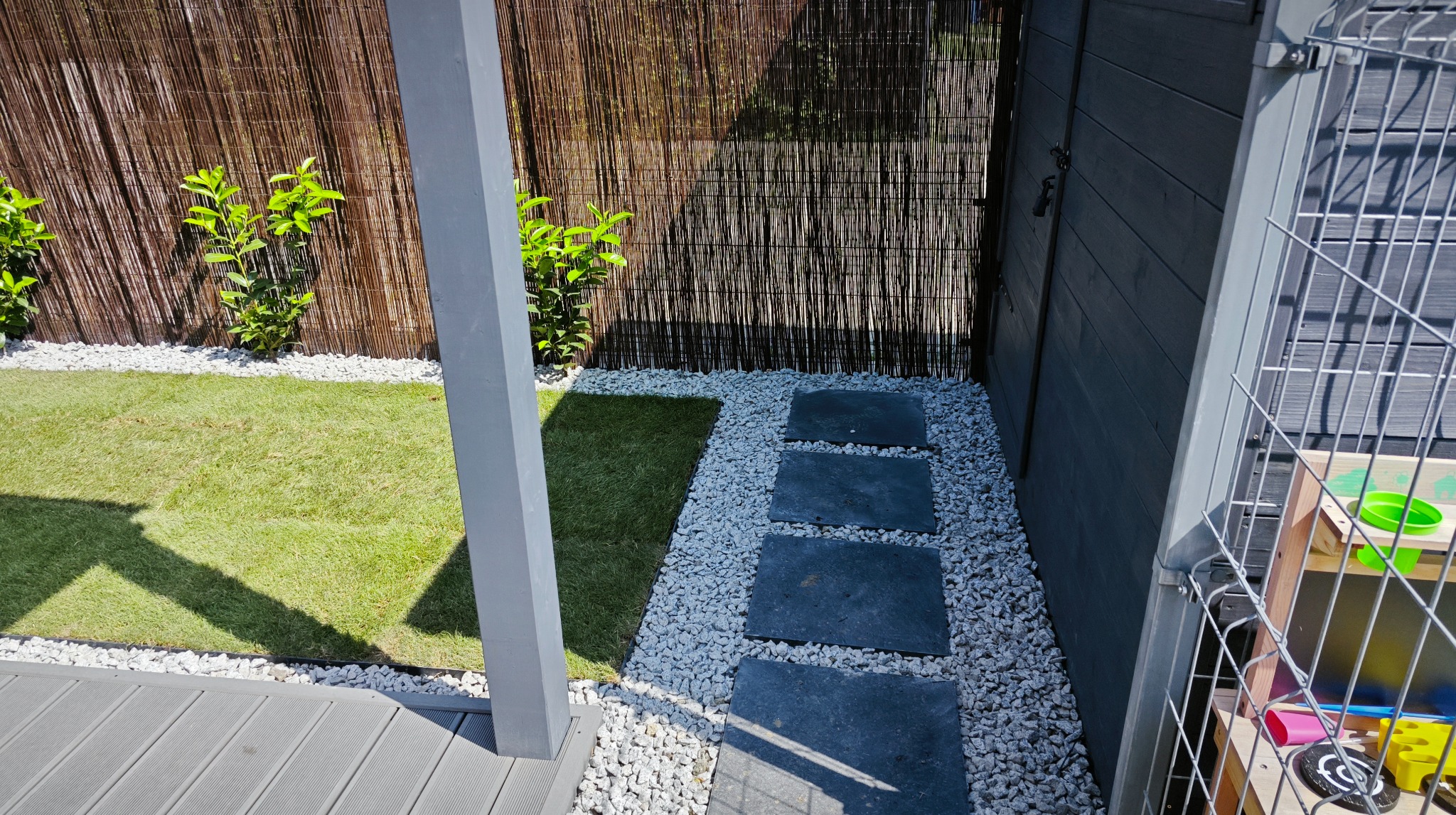 Aranżacja ogrodu: ścieżka z kamiennych płyt, białe kamyki, trawa i bambusowe ogrodzenie. Elementy drewniane w szarym odcieniu. Widok z góry.