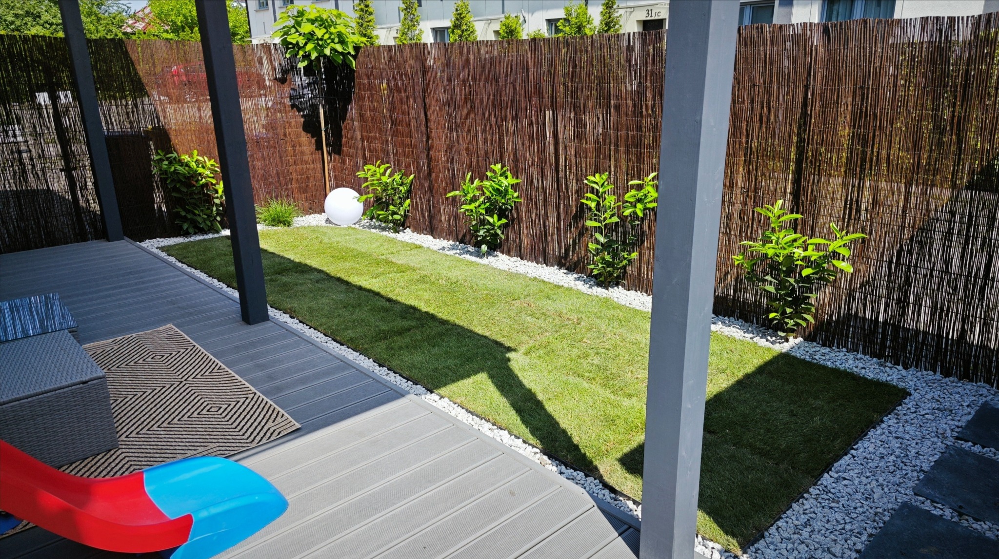 Zadbany ogród z trawą, ozdobnymi kamieniami i bambusowym ogrodzeniem. Widoczny fragment tarasu z meblami i zabawką.