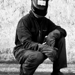 Czarno-białe zdjęcie spawacza w kombinezonie i masce ochronnej, w pozycji kucznej, z rękawicami roboczymi na kolanach. Tło stanowi surowa, teksturowana ściana.